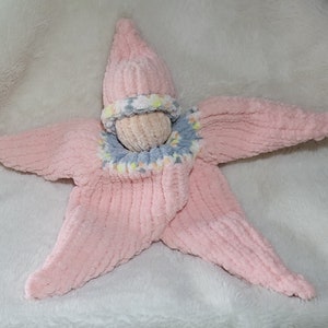 Könnte beinhalten: Ein rosa gestrickter Seestern mit einer grau-weißen Strickmütze. Der Seestern hat eine strukturierte Oberfläche und ist aus weichem Garn gefertigt.