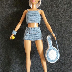 Könnte beinhalten: Ein gehäkeltes Puppenoutfit in Blau und Weiß, bestehend aus einem Crop-Top, Shorts, einer Mütze, einem Armband und einer Tasche mit einem runden Griff. Die Puppe trägt weiße Schuhe.