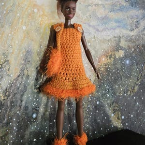 Könnte beinhalten: Puppe in einem orangefarbenen Häkelkleid mit passenden Stiefeln und einer Handtasche. Das Kleid hat einen flauschigen Saum und wird mit zwei Holzknöpfen verziert. Die Puppe hat dunkle Haut und lockiges Haar. Der Hintergrund ist ein farbenfrohes abstraktes Gemälde.