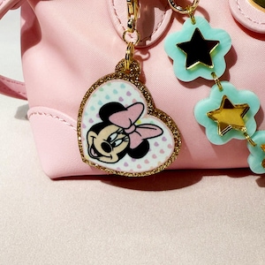 Puede incluir: Un bolso rosa con herrajes dorados y un charm de Minnie Mouse. El charm tiene forma de coraz&oacute;n con un borde dorado brillante y presenta a Minnie Mouse con un lazo rosa. Tambi&eacute;n hay un charm en forma de estrella en azul claro y dorado.