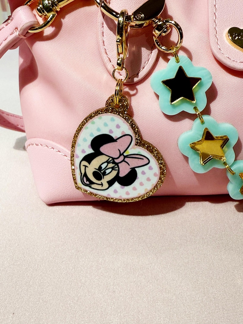 Puede incluir: Un bolso rosa con herrajes dorados y un charm de Minnie Mouse. El charm tiene forma de coraz&oacute;n con un borde dorado brillante y presenta a Minnie Mouse con un lazo rosa. Tambi&eacute;n hay un charm en forma de estrella en azul claro y dorado.