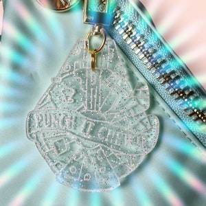 Puede incluir: Un llavero de plástico transparente con forma de Halcón Milenario de Star Wars con purpurina incrustada en el plástico. El llavero tiene el texto "Punch it Chewie" escrito en él.