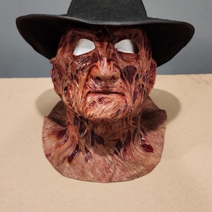 Puede incluir: Una máscara de cabeza completa con una tez texturizada, cicatrizada y de color marrón rojizo con surcos profundos y detalles realistas de la piel. La máscara tiene cuencas oculares blancas y está rematada con un sombrero fedora de fieltro negro.