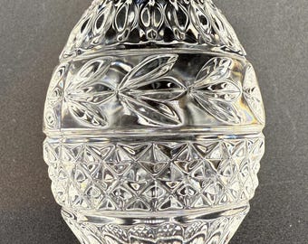 May include: A clear, decorative egg-shaped container with a lid. The glass is intricately cut with floral and geometric patterns. The egg is sitting on a dark surface.