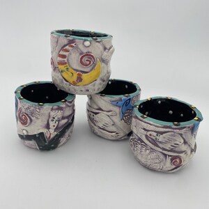 May include: Four ceramic cups with whimsical designs. Each cup features a different design, including a crescent moon, a bird, a woman's face, and a swirling pattern. The cups are all white with a teal rim and black accents.