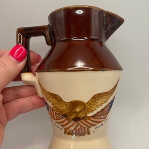 May include: A brown ceramic creamer with a white base. It features a golden eagle with red, white, and blue stripes, and the text "The Spirit of" and "America" on either side of the eagle.