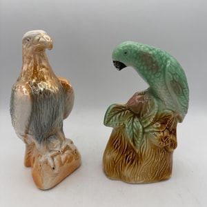 May include: Two ceramic bird figurines. One is an eagle with a white head, gray body, and orange accents. The other is a green parrot perched on a brown base with green leaves. Both are detailed and decorative.