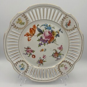 May include: A white porcelain plate with a delicate openwork design and a floral pattern in shades of pink, red, yellow, and blue. The plate has a scalloped edge and is decorated with gold accents.