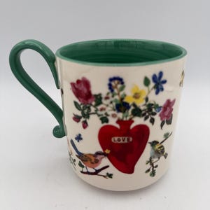 May include: Cream-colored ceramic mug with a green handle and rim. The mug features a red heart-shaped vase with the word "LOVE" and a bouquet of colorful flowers. Birds perch on branches around the heart.