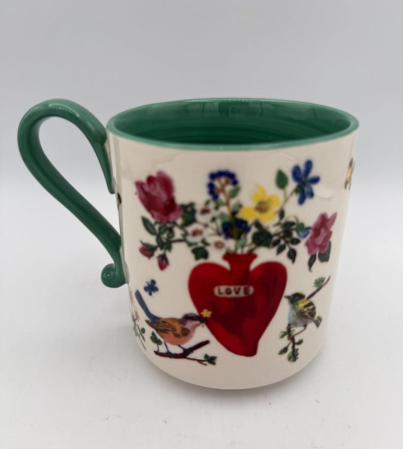 May include: Cream-colored ceramic mug with a green handle and rim. The mug features a red heart-shaped vase with the word "LOVE" and a bouquet of colorful flowers. Birds perch on branches around the heart.