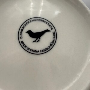 May include: Close-up of the bottom of a white ceramic bowl. A black circular emblem features a bird silhouette and the text "Dishwasher & Microwave Safe" and "Made in China Fabriqu&eacute; en Chine".