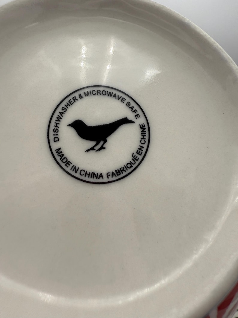 May include: Close-up of the bottom of a white ceramic bowl. A black circular emblem features a bird silhouette and the text "Dishwasher & Microwave Safe" and "Made in China Fabriqu&eacute; en Chine".