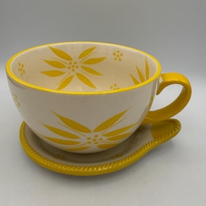 May include: Cream-colored oversized teacup and saucer set with yellow floral designs. The cup has a large handle and sits on a matching saucer. The rim and handle are bright yellow. A decorative kitchenware item.