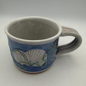 May include: A ceramic mug featuring a light blue glazed exterior with a raised, white, textured seashell relief design. The mug has a wide, sturdy handle with three distinct ridges and a smooth, off-white interior finish with a visible rim.