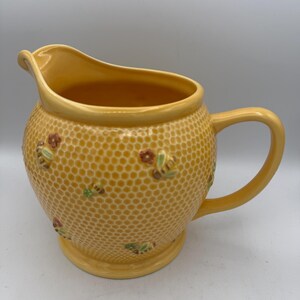 May include: A yellow ceramic pitcher featuring a raised honeycomb texture across the exterior surface. The pitcher is decorated with small, colorful, three-dimensional bees and flowers scattered across the hexagonal pattern.