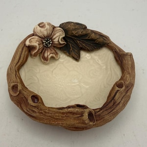 May include: A round ceramic dish with a cream-colored interior featuring an embossed floral pattern. The rim is textured to resemble brown tree bark and is decorated with a sculpted cream flower and dark brown leaves.