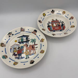 May include: Two white ceramic plates with colorful illustrations. One plate features a scene with a camel, people, and a building. The other plate is a bowl with a red bus illustration. Both plates have a blue dotted border with cartoon faces.