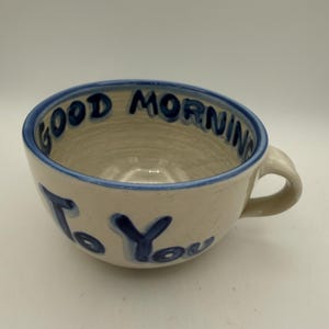 May include: A ceramic coffee mug with a blue rim and handle. The words "GOOD MORNING To You" are written in blue on the mug's exterior. The mug has a beige interior.