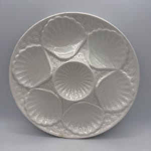 May include: A white ceramic oyster serving plate. The plate features six shell-shaped compartments around a central, smaller, circular compartment. The plate has a textured, ridged design around the edges.