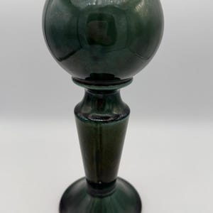 May include: A dark green ceramic vase with a spherical top and a pedestal base. The vase has a glossy finish and a classic, elegant design. The vase is approximately 10 inches tall.