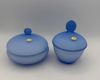 May include: Two frosted blue glass jars with matching lids, each featuring a small spherical knob handle and a decorative gold-colored emblem on the lid. The jars have a smooth, matte finish and a rounded, bulbous shape for storage.