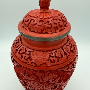 May include: A red, lidded jar with intricate carved floral designs. The jar has a rounded body and a lid with a knob. The carvings are detailed and cover the entire surface of the jar, creating a textured appearance. The jar is a vibrant shade of red.