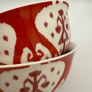May include: Two ceramic bowls stacked, featuring a bold red and white pattern. The bowls have a glossy finish and a unique design, with a white interior and a red exterior adorned with white and maroon shapes. The bowls are ideal for serving food.