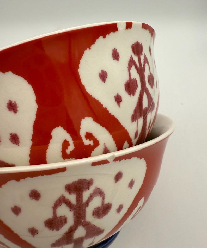 May include: Two ceramic bowls stacked, featuring a bold red and white pattern. The bowls have a glossy finish and a unique design, with a white interior and a red exterior adorned with white and maroon shapes. The bowls are ideal for serving food.