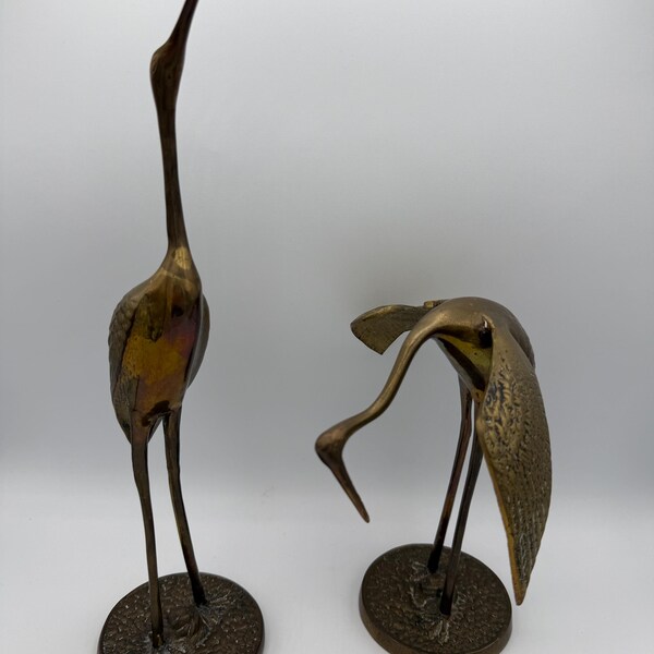 Peut inclure: Deux sculptures de grues en bronze. Une grue se dresse, le cou tendu, tandis que l'autre est courbée. Les deux sont montées sur des socles circulaires. Les sculptures ont une surface texturée et une finition métallique chaude.