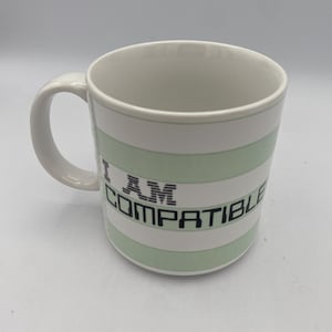 May include: White ceramic mug with a curved handle. The mug features horizontal stripes in white and light green. The text "I AM COMPATIBLE" is printed in black on the front of the mug.