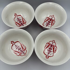 May include: Four white ceramic bowls with a red and maroon decorative pattern inside. The bowls have a simple, rounded shape and a glossy finish. The red design is a stylized floral or abstract motif.