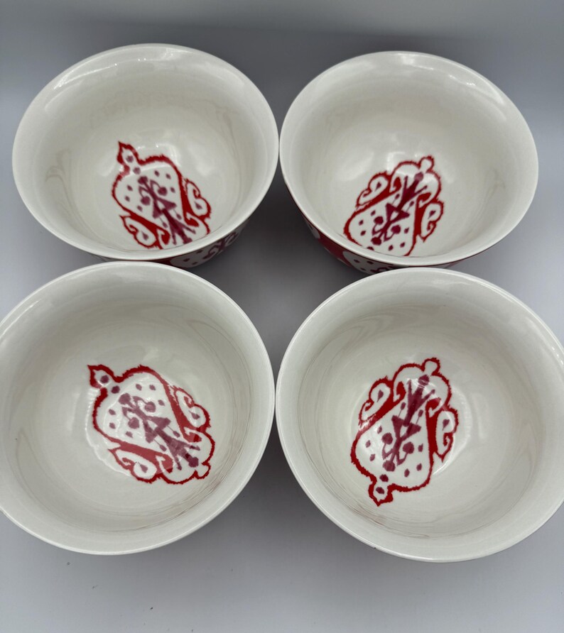 May include: Four white ceramic bowls with a red and maroon decorative pattern inside. The bowls have a simple, rounded shape and a glossy finish. The red design is a stylized floral or abstract motif.