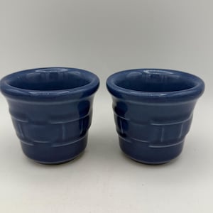 May include: Two navy blue ceramic cups with a textured, brick-like pattern. The cups have a rounded top and a slightly flared base. The cups are displayed on a white surface.