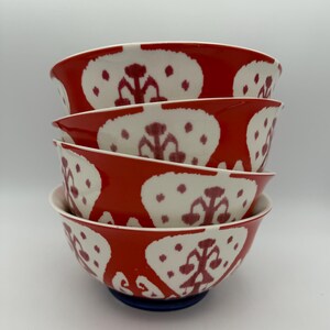 May include: A stack of four ceramic bowls. The bowls have a vibrant red exterior with a white, patterned design. The interior of the bowls is white. The bowls are stacked on top of each other, with a blue base supporting the bottom bowl.