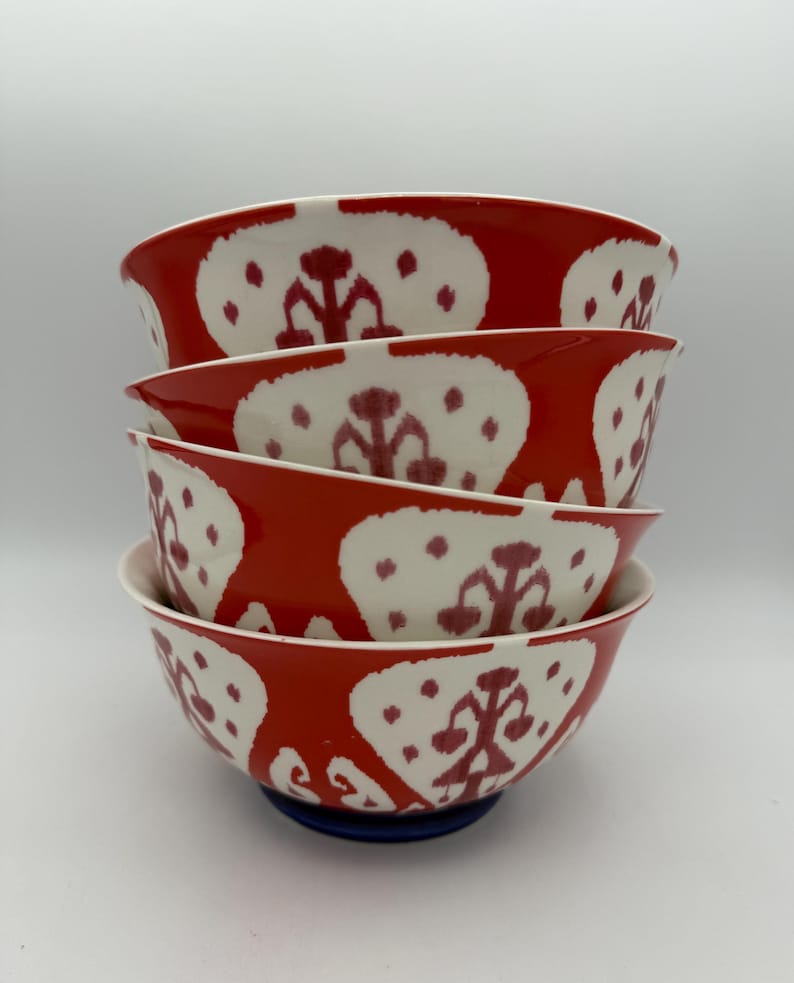 May include: A stack of four ceramic bowls. The bowls have a vibrant red exterior with a white, patterned design. The interior of the bowls is white. The bowls are stacked on top of each other, with a blue base supporting the bottom bowl.