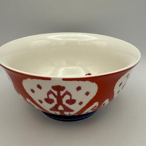 May include: A ceramic bowl with a white interior and a red exterior decorated with white, stylized floral patterns. The bowl sits on a dark blue base. This decorative bowl is suitable for serving food.