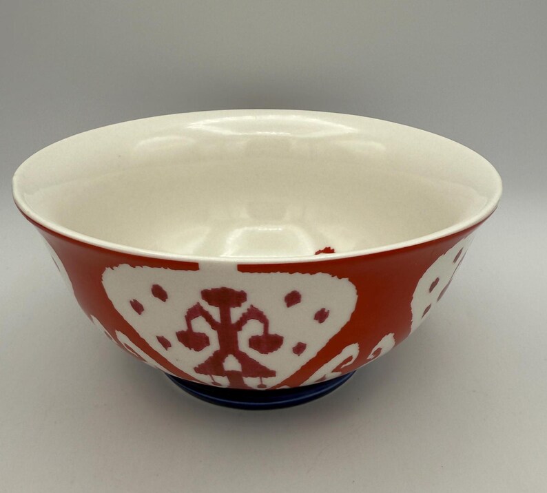 May include: A ceramic bowl with a white interior and a red exterior decorated with white, stylized floral patterns. The bowl sits on a dark blue base. This decorative bowl is suitable for serving food.