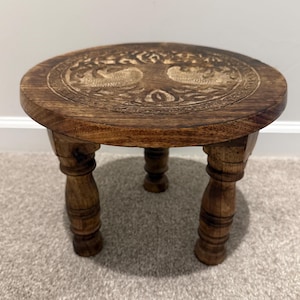 May include: A small, round, dark brown wooden stool featuring three carved legs and a circular top surface. The top of the stool displays an intricate, engraved design of a tree with sprawling branches and roots, centered within a decorative border.