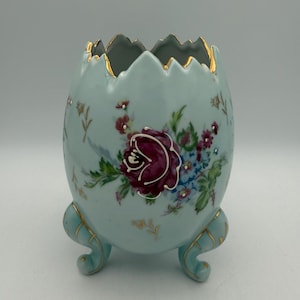 May include: A light blue, egg-shaped ceramic vase with a floral design. The vase has a scalloped rim with gold trim and three curved feet. The floral design features a large burgundy rose and smaller flowers.