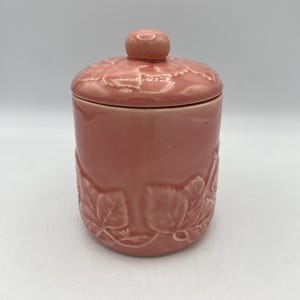 May include: A pink ceramic jar with a lid featuring a floral design. The jar has a smooth, glossy finish and is decorated with raised leaves.