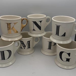 May include: A collection of six cream-colored ceramic mugs with handles, each featuring a single large letter on the front. The letters displayed are K, N, L, D, M, P, and G. The letter K is gold, while the others are black with various patterns.