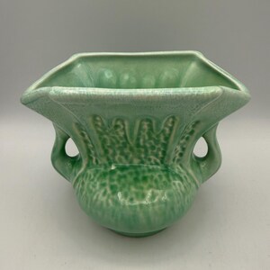 May include: A light green ceramic vase with a textured surface and two handles. The vase has a square top and a rounded base. The design includes a speckled pattern and decorative elements along the sides. The vase is approximately 7 inches wide.