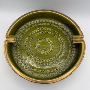May include: A vintage green ceramic ashtray with a gold rim and accents. The ashtray features a circular design with heart and dot patterns. It has two built-in rests for cigarettes.