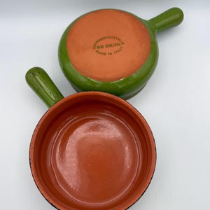 May include: Two small, round, terracotta-colored ceramic pans with green handles and rims. One pan is upside down, revealing a terracotta interior and the text "FABBRICA IN ITALIA DE SILVA". The other pan is right-side up.