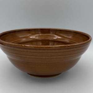 May include: A handmade, brown ceramic bowl with a wide, shallow design. The bowl has a smooth, glossy finish and visible horizontal ridges. The color is a warm, earthy tone, suitable for serving food or as a decorative piece.