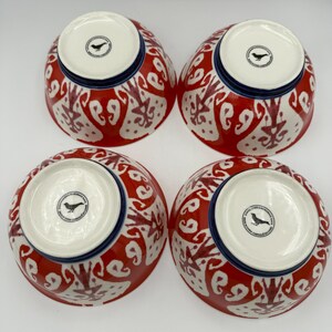 May include: Four ceramic bowls with a red and white patterned design. The bowls have a blue rim and a white interior. Each bowl has a black bird logo on the bottom. The bowls are arranged in a square.
