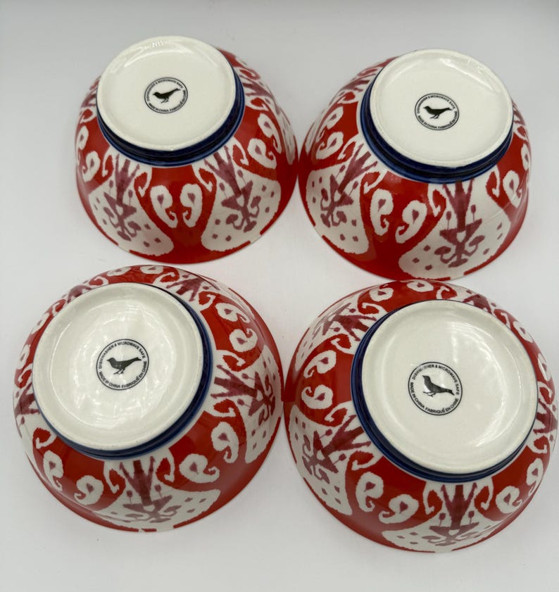 May include: Four ceramic bowls with a red and white patterned design. The bowls have a blue rim and a white interior. Each bowl has a black bird logo on the bottom. The bowls are arranged in a square.