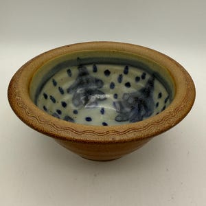 May include: A handmade ceramic bowl with a light brown exterior and a blue and white interior. The rim has a textured, wavy design. The interior features blue abstract shapes and dots. A unique piece for serving or display.