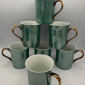 May include: A stack of eight teal-colored ceramic mugs with gold-colored handles. The mugs have a speckled finish and a white interior. The handles are curved and have a metallic gold finish. The mugs are arranged in a pyramid shape.