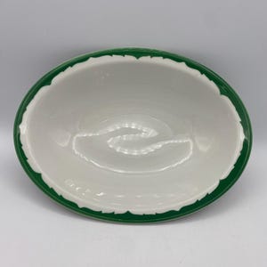 May include: White oval ceramic bowl with a scalloped green rim. The bowl has a smooth, glossy finish and is empty. The green rim adds a decorative touch to the otherwise plain white bowl. This bowl is suitable for serving food.